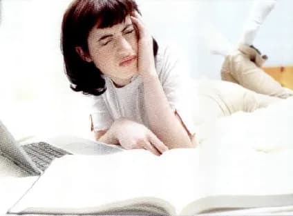 A young woman lying on a bed with an open book and a laptop, holding her head as if she is tired and under pressure.