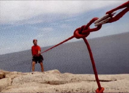 A climber hanging on a red rope at the edge of a high rock above the sea.
