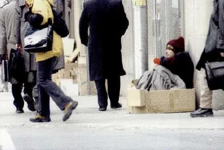 A homeless person sitting on a pavement among people who walk past.