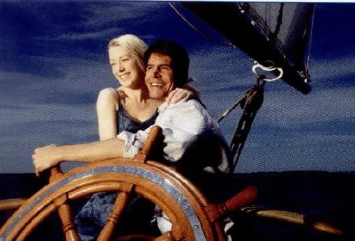 A smiling young couple on a sailing boat, with the man steering at the wheel.