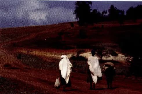 A few people in white clothes walking along a dry red dirt road in a poor, empty landscape.