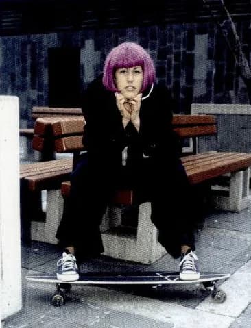 A young woman with pink hair and black clothes, sitting on a bench with a skateboard.